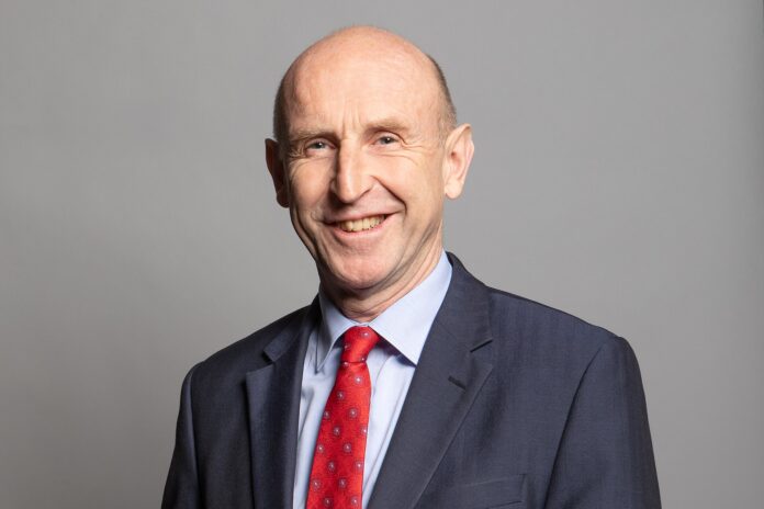 Portrett av John Healey i dress og rødt slips mot lys bakgrunn.