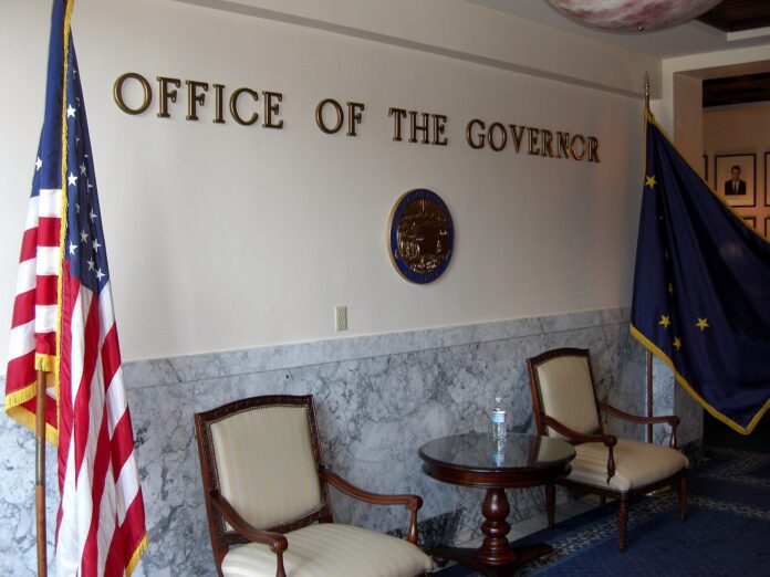 Interiør fra guvernørkontoret i Alaska State Capitol, med skiltet «Office of the Governor», to stoler, et lite bord og amerikansk og alaskisk flagg.