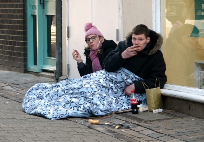 To personer sitter på fortauet ved en butikkfasade i Norwich, delvis dekket av et teppe.